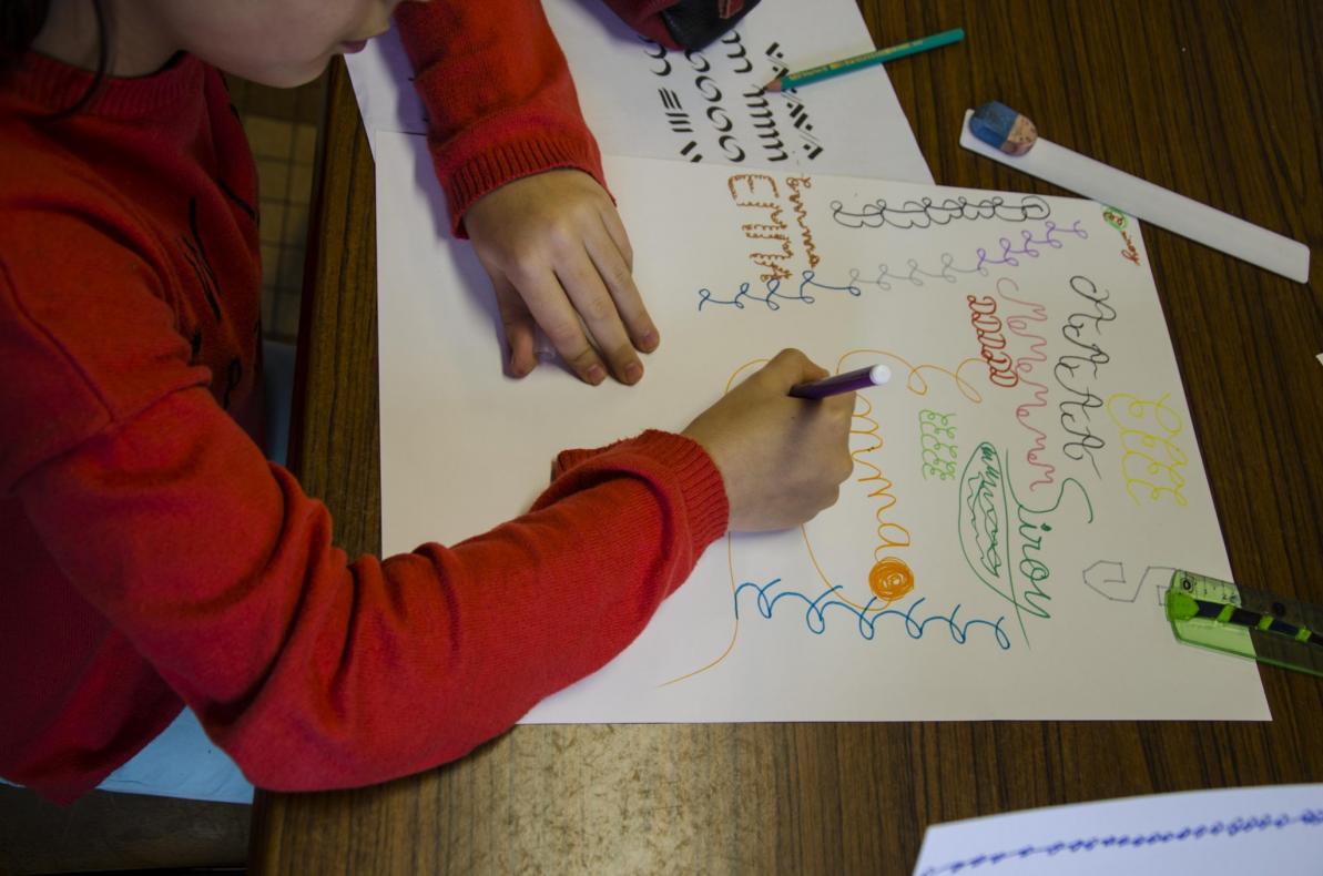 Élève en train de faire la composition contenant sa signature et des traits calligraphiques de base utilisés en tant que motifs d’ornementation. À gauche, document distribué contenant ces exercices. Outil utilisé : feutres monolinéaires en plusieurs couleurs.