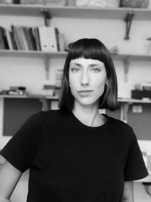 Portrait en noir et blanc d'une personne aux cheveux mi-longs. On distingue en fond des étagères d'une bibliothèque.